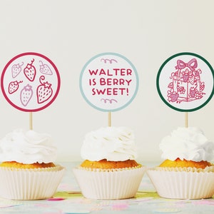 May include: Three cupcakes with white frosting and red and green circular toppers. The toppers have illustrations of strawberries and a gift box with a bow. The text on one topper reads "Walter is berry sweet!"