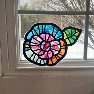 May include: A colourful paper flower with a pink centre and blue, green, and orange petals. The flower is attached to a green leaf and is displayed on a window.
