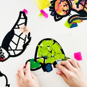May include: A child's hand placing green tissue paper onto a black paper turtle outline. The turtle is being decorated with green, blue, and white tissue paper.