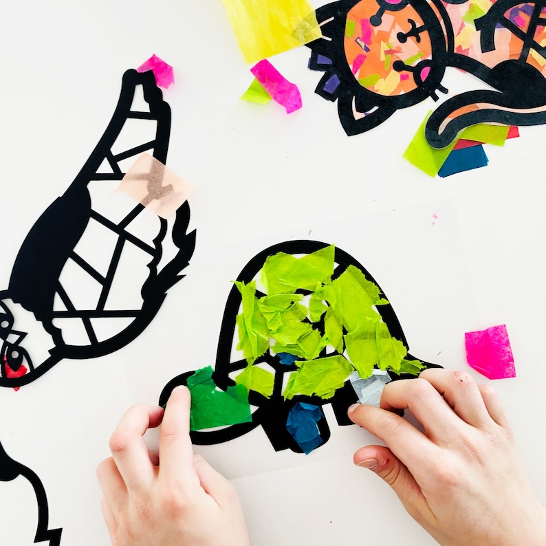 May include: A child's hand placing green tissue paper onto a black paper turtle outline. The turtle is being decorated with green, blue, and white tissue paper.