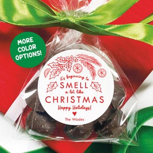 May include: A clear cellophane bag filled with dark chocolate candies, tied with a green ribbon, and set against a red background. A white circular sticker with red text reads "It's beginning to smell a lot like Christmas".