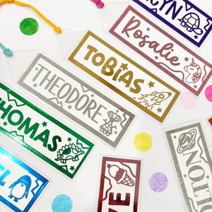 May include: A collection of personalized name bookmarks in various colors. Each bookmark features a name in a decorative font, with accompanying illustrations like a rocket, robot, or unicorn. The bookmarks have colorful tassels and are scattered with confetti.