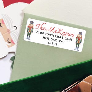 May include: A white rectangular label with the text "The McKnews" in red script, along with an address. The label is decorated with illustrations of nutcrackers in festive attire. The label is placed on a light green envelope.