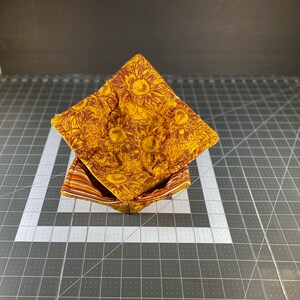 May include: A set of two brown and yellow fabric bowl cozies with a sunflower pattern. The cozies are folded and stacked on top of each other.