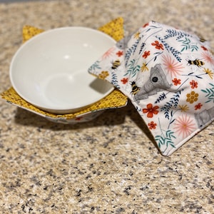 May include: A white ceramic bowl with a floral print bowl cozy. The cozy has a white background with yellow, red, and pink flowers, bees, and small animals.