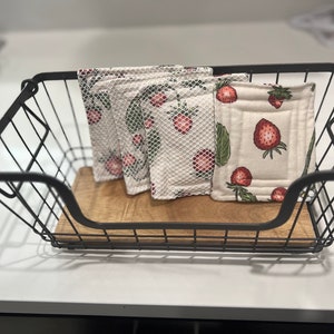 May include: Four white cotton dishcloths with a strawberry pattern are stacked in a black wire basket. The dishcloths are made of a soft, absorbent material and are perfect for cleaning up spills and messes in the kitchen.