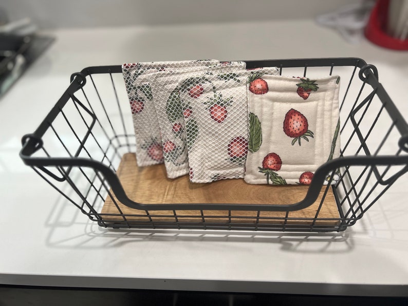 May include: Four white cotton dishcloths with a strawberry pattern are stacked in a black wire basket. The dishcloths are made of a soft, absorbent material and are perfect for cleaning up spills and messes in the kitchen.