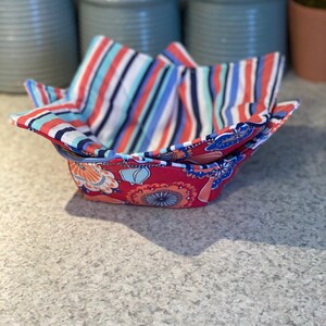 May include: A set of two fabric bowls, one with a red, orange, and blue floral pattern and the other with blue, red, and white stripes. The bowls are folded and ready to be used.