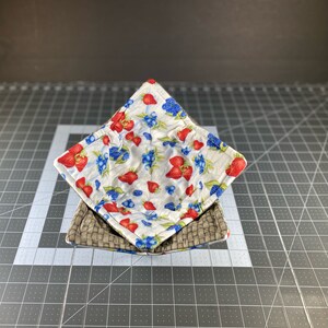 May include: A quilted bowl cozy with a white background and a pattern of red strawberries and blue blueberries. The cozy is folded in half and is sitting on a gray cutting mat.