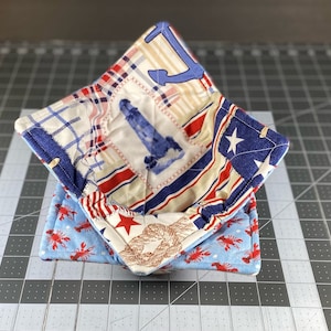 May include: A set of two fabric bowl cozies. The top cozy is made of a patchwork fabric with red, white, and blue stripes, a lighthouse, and a star. The bottom cozy is blue with red lobsters and a white rope design.