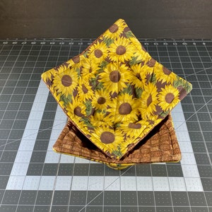 May include: Two square fabric bowl cozies. The top cozy features a yellow sunflower pattern on a brown background. The bottom cozy has a brown woven pattern. These cozies are designed to be used in the microwave.