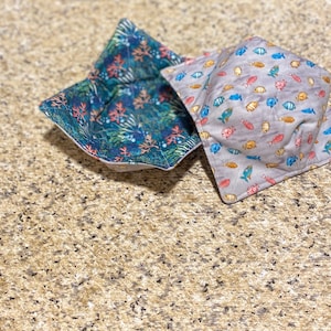 May include: Two square fabric bowl cozies. One features a teal and coral reef pattern. The other has a gray background with colorful fish illustrations. These cozies are designed to be placed in the microwave to heat up food.