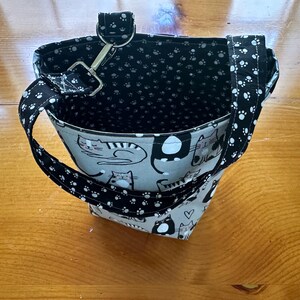 May include: A small, open tote bag with a cat pattern on the exterior. The bag features a black and white paw print pattern on the handles and interior lining. A metal clasp and ring secure the handle to the bag.