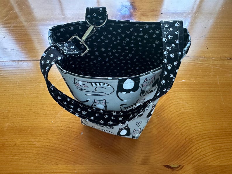 May include: A small, open tote bag with a cat pattern on the exterior. The bag features a black and white paw print pattern on the handles and interior lining. A metal clasp and ring secure the handle to the bag.