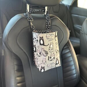 May include: A gray car trash bag with a cat pattern hangs from a car seat headrest. The bag features various cat illustrations in black and white on a gray background. The straps are black with white polka dots.