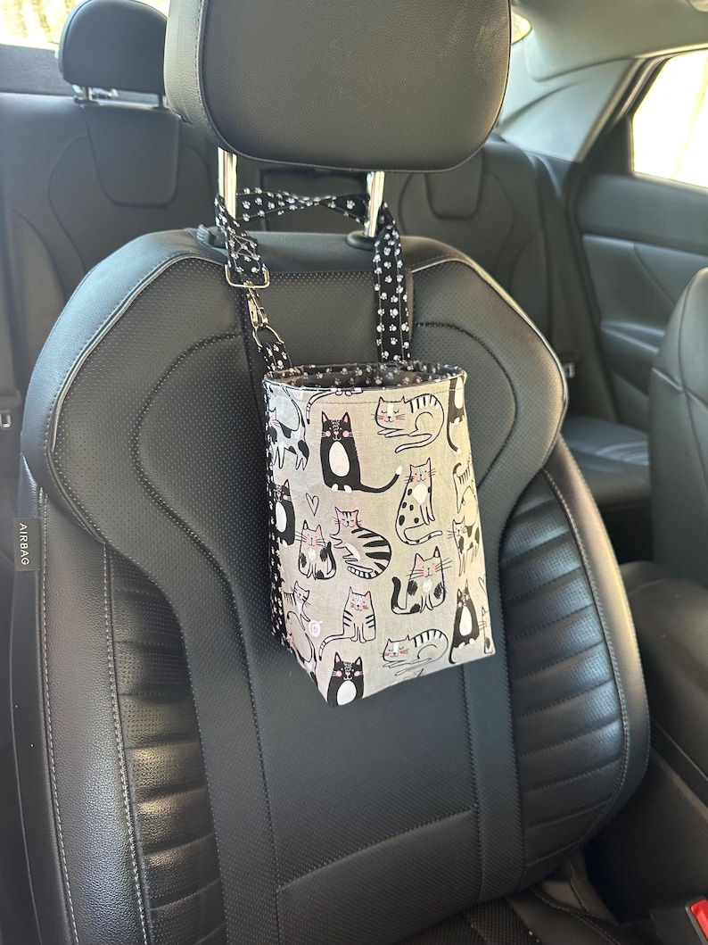 May include: A gray car trash bag with a cat pattern hangs from a car seat headrest. The bag features various cat illustrations in black and white on a gray background. The straps are black with white polka dots.