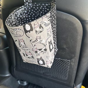 May include: A car seat organizer with a cat pattern. The organizer is made of fabric and hangs from the headrest. The main fabric is gray with black and white cat illustrations. The interior and straps feature a black and white paw print design.