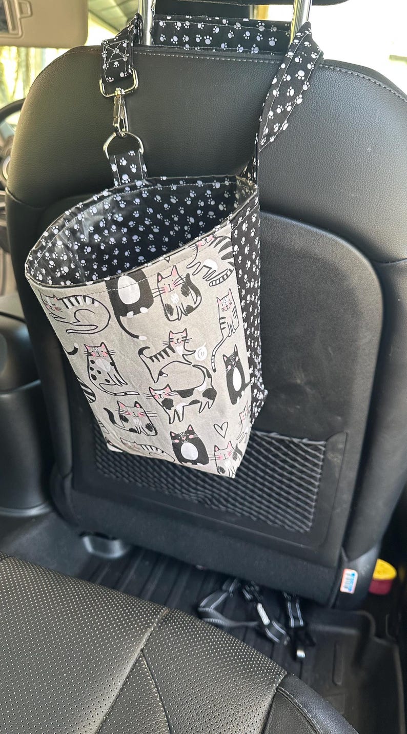 May include: A car seat organizer with a cat pattern. The organizer is made of fabric and hangs from the headrest. The main fabric is gray with black and white cat illustrations. The interior and straps feature a black and white paw print design.