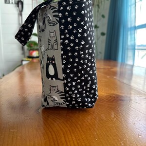 May include: A cylindrical fabric bag with a handle, featuring a cat-themed design. One side displays various cat illustrations on a gray background, while the other side is covered in a black and white paw print pattern. The handle also has a paw print design.