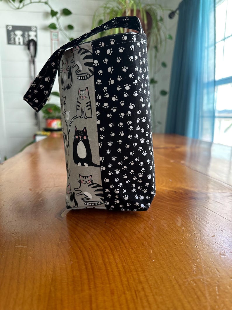 May include: A cylindrical fabric bag with a handle, featuring a cat-themed design. One side displays various cat illustrations on a gray background, while the other side is covered in a black and white paw print pattern. The handle also has a paw print design.