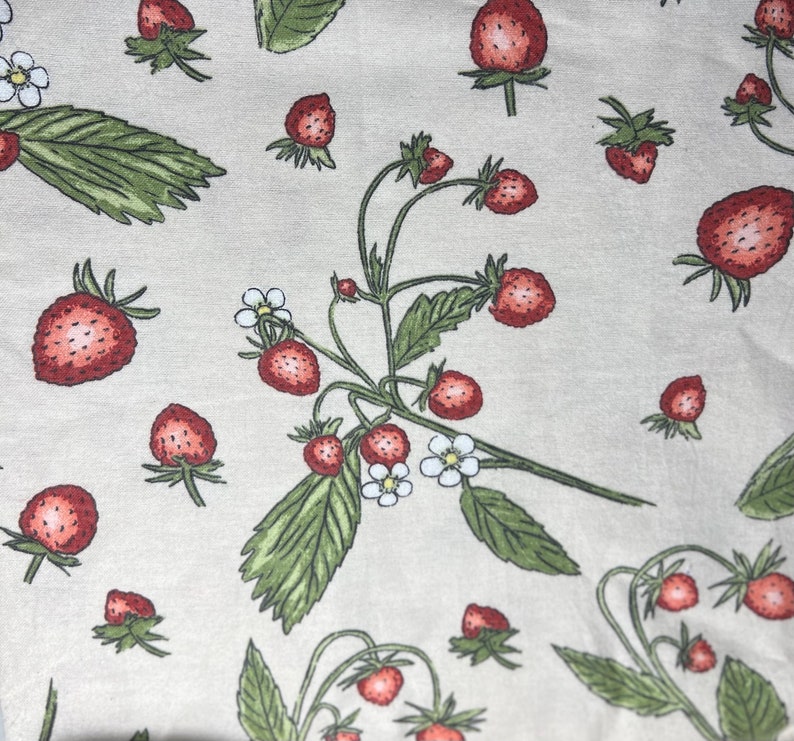 May include: A beige fabric with a repeating pattern of red strawberries and green leaves. The strawberries are in various stages of ripeness and some have white flowers.