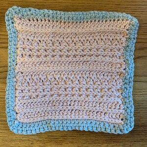 Könnte beinhalten: Ein quadratisches, handgefertigtes gehäkeltes Waschtuch. Die Mitte ist hellpfirsichfarben, mit einem strukturierten Stichmuster. Der Rand ist hellblau, mit einem gewellten Rand. Das Waschtuch liegt auf einer Holzoberfläche.