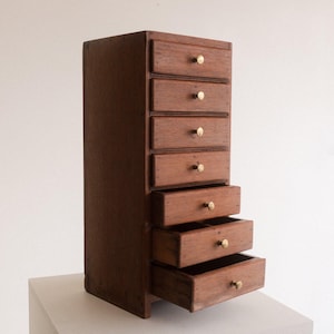 May include: A small, wooden chest of drawers with seven drawers, some of which are open. The chest is made of dark brown wood and has brass knobs. It is displayed on a white surface.