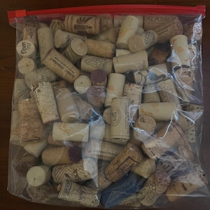 May include: A clear plastic bag filled with assorted used wine corks. The corks are cylindrical, made of natural cork, and feature printed text and designs from various wineries. The bag is sealed with a red zipper.
