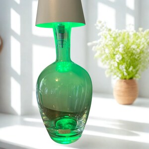 May include: A green glass bottle lamp with a beige lampshade. The lamp has a unique design, with a bottle-shaped base and a slender neck. The lamp emits a soft green glow, creating a warm and inviting ambiance. The lamp is a decorative item.
