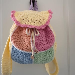 May include: A handmade crochet backpack in pastel colors, including yellow, pink, peach, blue, and green. The bag features a scalloped flap and drawstring closure. The straps and handle are yellow. A wooden star charm hangs from the drawstring.
