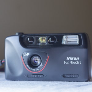 Pode incluir: Câmara de filme Nikon Fun-Touch 2 preta de 35 mm. A câmara tem um flash embutido e uma lente de 34 mm com uma abertura de 1:4,5. A lente tem um detalhe vermelho e o texto "Nikon Lens".
