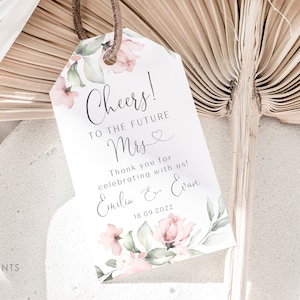 May include: A white tag with floral designs in pink and green. The tag says "Cheers! To the future Mrs." with a heart symbol. It also says "Thank you for celebrating with us! Emilia & Evan 18.09.2022".