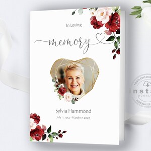 May include: A white card with a gold heart frame and a photo of a woman inside. The heart frame is decorated with red and white flowers. The text "In Loving Memory" is written in cursive at the top of the card. The text "Sylvia Hammond" is written below the photo, followed by the dates "July 11, 1952 - March 17, 2020".