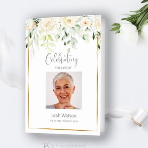May include: A white memorial booklet with a gold border and a floral design. The booklet has a photo of a woman and the text "Celebrating the life of Leah Watson July 15, 1962 - March 11, 2020".