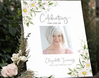 May include: A white sign on an easel with floral and botanical accents. The sign reads "Celebrating the Life of Elizabeth Jennings" with a photo and dates July 15, 1949 - February 11, 2022. White daisies and green leaves decorate the edges.