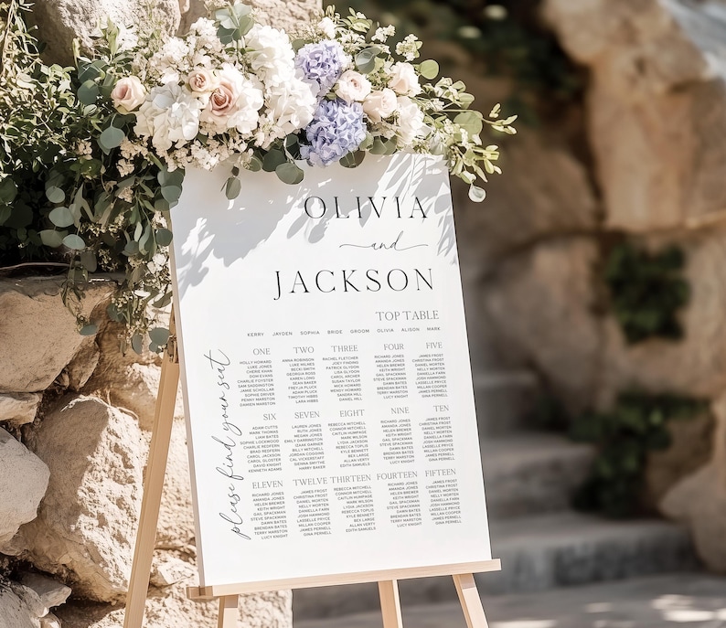 May include: A white wedding seating chart on an easel, adorned with floral arrangements of white, pink, and purple flowers. The chart displays names under the headings "OLIVIA and JACKSON" and "TOP TABLE."