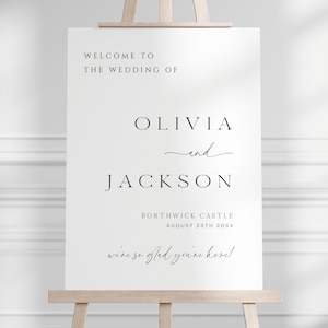 Op de afbeelding: Een wit trouwbord op een houten ezel. De zwarte tekst luidt "WELCOME TO THE WEDDING OF OLIVIA and JACKSON", met de trouwdatum en locatie. De tekst bevat ook de zin "we're so glad you're here!"