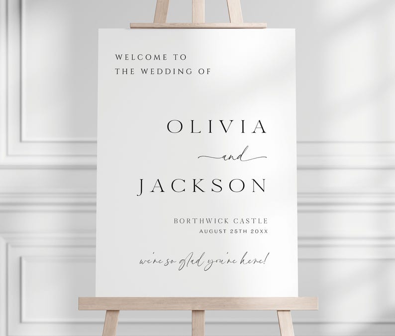Op de afbeelding: Een wit trouwbord op een houten ezel. De zwarte tekst luidt "WELCOME TO THE WEDDING OF OLIVIA and JACKSON", met de trouwdatum en locatie. De tekst bevat ook de zin "we're so glad you're here!"