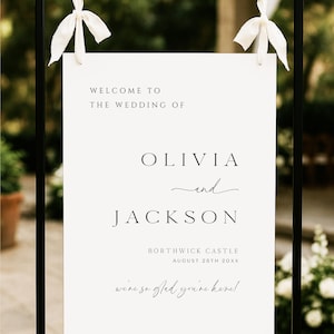 Op de afbeelding: Een welkomstbord voor een bruiloft met de tekst "WELCOME TO THE WEDDING OF OLIVIA and JACKSON" in zwarte letters. Het bord bevat ook de locatie en datum: BORTHWICK CASTLE, 25 AUGUSTUS 20XX. Het bord hangt aan een zwart metalen frame met witte linten.