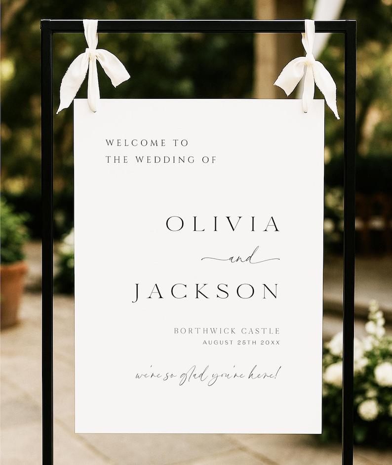 Op de afbeelding: Een welkomstbord voor een bruiloft met de tekst "WELCOME TO THE WEDDING OF OLIVIA and JACKSON" in zwarte letters. Het bord bevat ook de locatie en datum: BORTHWICK CASTLE, 25 AUGUSTUS 20XX. Het bord hangt aan een zwart metalen frame met witte linten.
