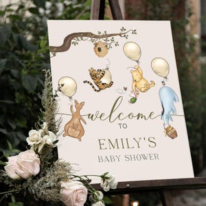 May include: A cream-coloured baby shower sign with illustrations of Winnie the Pooh characters and balloons. The sign reads "welcome to EMILY'S BABY SHOWER" in a decorative font. A floral arrangement with pink and white roses is in the foreground.