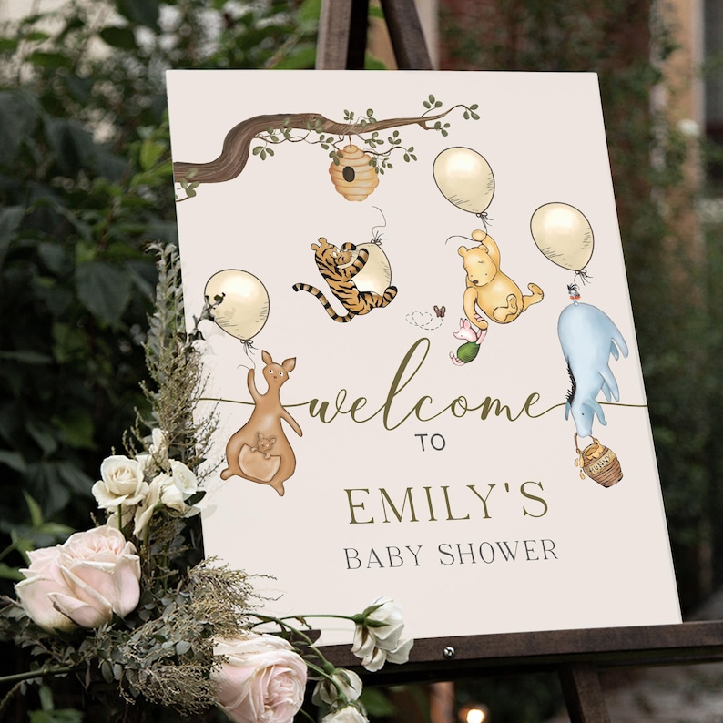 May include: A cream-coloured baby shower sign with illustrations of Winnie the Pooh characters and balloons. The sign reads "welcome to EMILY'S BABY SHOWER" in a decorative font. A floral arrangement with pink and white roses is in the foreground.