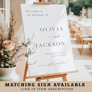 Op de afbeelding: Een wit welkomstbord voor een bruiloft op een houten ezel met zwarte tekst: WELCOME TO THE WEDDING OF OLIVIA and JACKSON BORTHWICK CASTLE AUGUST 25TH 20XX we're so glad you're here! Een vaas met een roze roos en gedroogd gras staat ernaast.