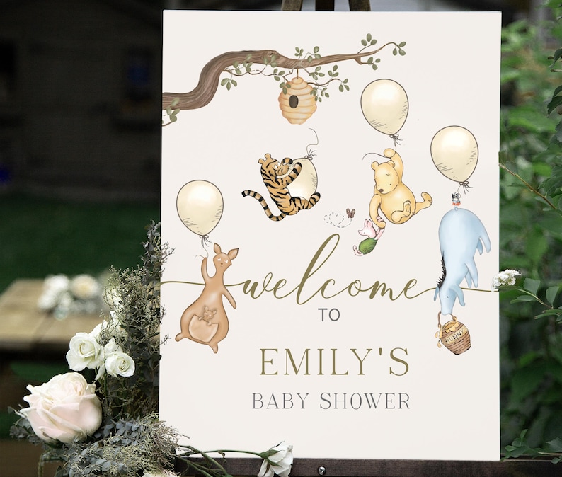 May include: A cream-coloured baby shower sign with illustrations of Winnie the Pooh characters. The sign includes the text "welcome to Emily's baby shower" in gold script, with balloons and a beehive.