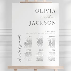 May include: A white wedding seating chart on an easel. The names "Olivia and Jackson" are at the top, with guest names organized by table number below. The words "please find your seat" are written on the left side.