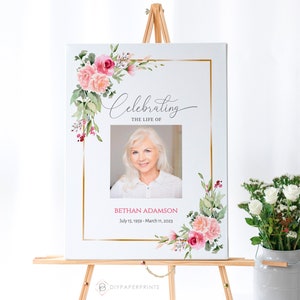 May include: A white canvas sign with a gold frame and pink floral accents. The sign reads "Celebrating the Life of Bethan Adamson July 15, 1959 - March 11, 2023". A photo of a woman is in the center of the sign.