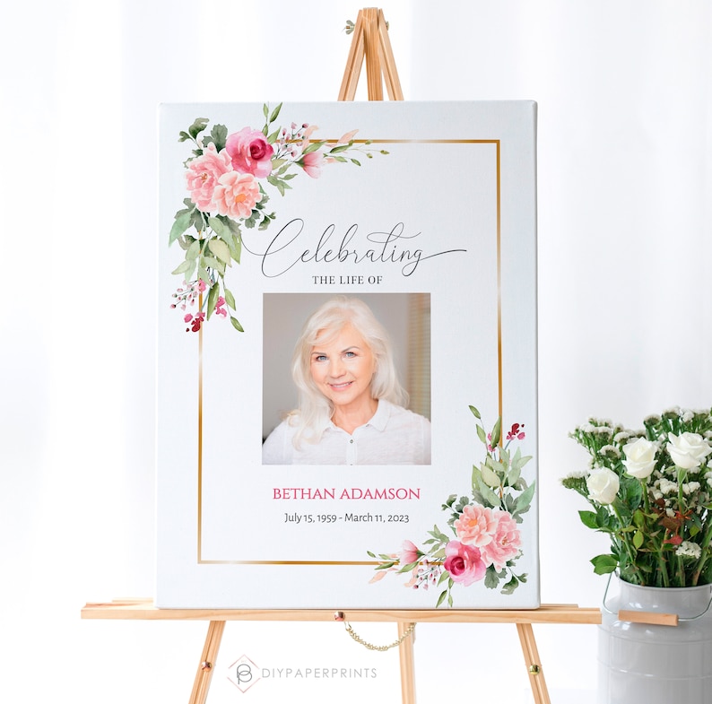 May include: A white canvas sign with a gold frame and pink floral accents. The sign reads "Celebrating the Life of Bethan Adamson July 15, 1959 - March 11, 2023". A photo of a woman is in the center of the sign.