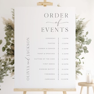 Op de afbeelding: Een wit trouwevenementbord op een houten ezel, met de tekst "ORDER of EVENTS" in elegant zwart schrift. Het bord geeft het schema weer, inclusief de ceremonie, het diner en het vuurwerk, met tijden. De namen "OLIVIA and JACKSON" worden ook weergegeven.