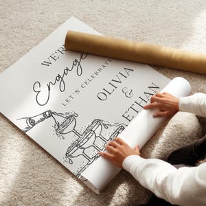 K&ouml;nnte beinhalten: Ein wei&szlig;es Schild mit schwarzem Text, auf dem steht "We're Engaged, Let's Celebrate Olivia & Ethan" mit einer Illustration einer Champagnerflasche und Gl&auml;sern. Ein aufgerolltes Poster und eine braune R&ouml;hre sind ebenfalls im Bild.