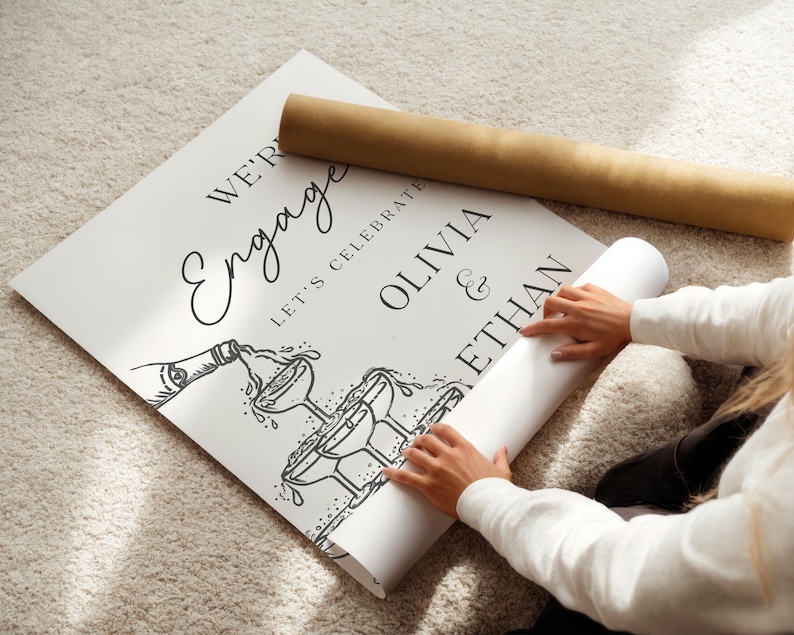 K&ouml;nnte beinhalten: Ein wei&szlig;es Schild mit schwarzem Text, auf dem steht "We're Engaged, Let's Celebrate Olivia & Ethan" mit einer Illustration einer Champagnerflasche und Gl&auml;sern. Ein aufgerolltes Poster und eine braune R&ouml;hre sind ebenfalls im Bild.